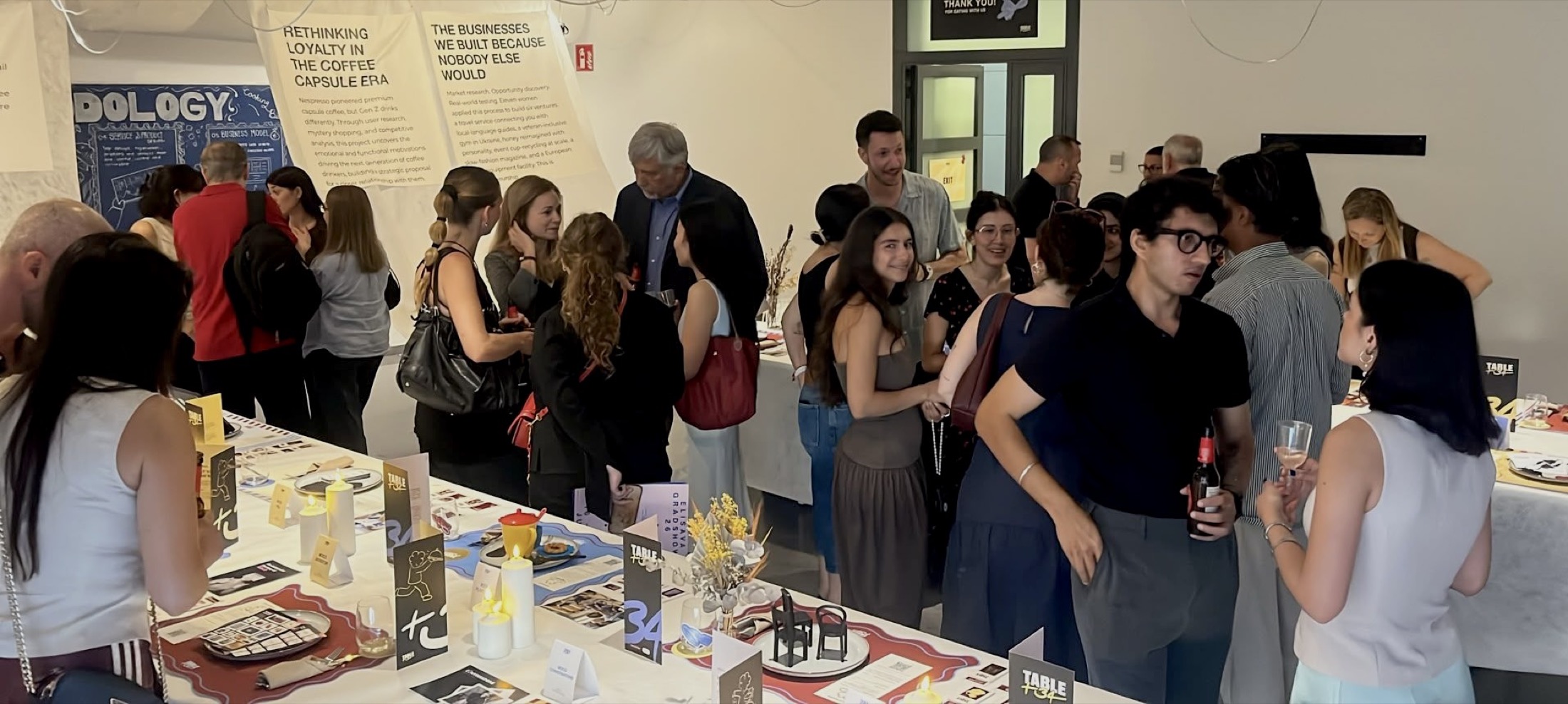 Guests filling the room at the Table+34 Grad Show, talking around the tables